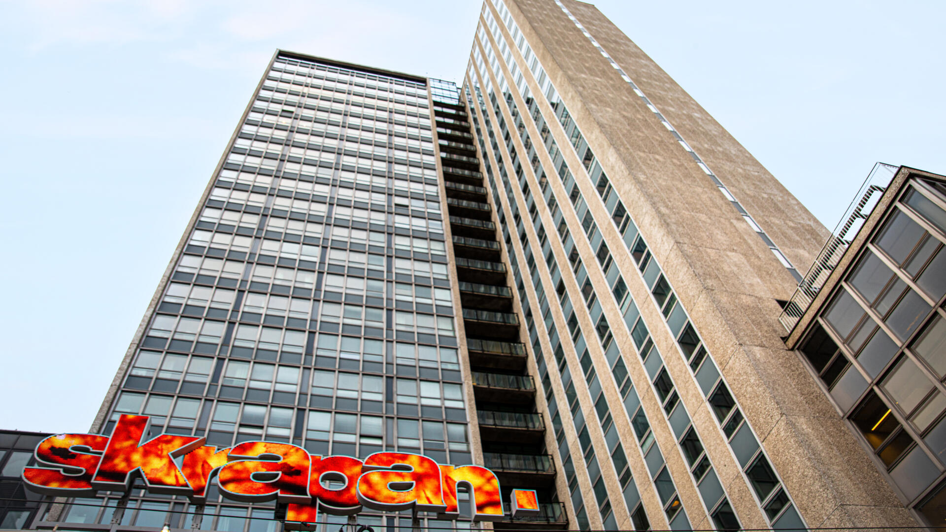 Den betong- och glasförsedda fasaden av gamla skatteskrapan sträcker sig upp mot en blå himmel. Längst ner syns den färgsprakande skylten som lyder "skrapan".