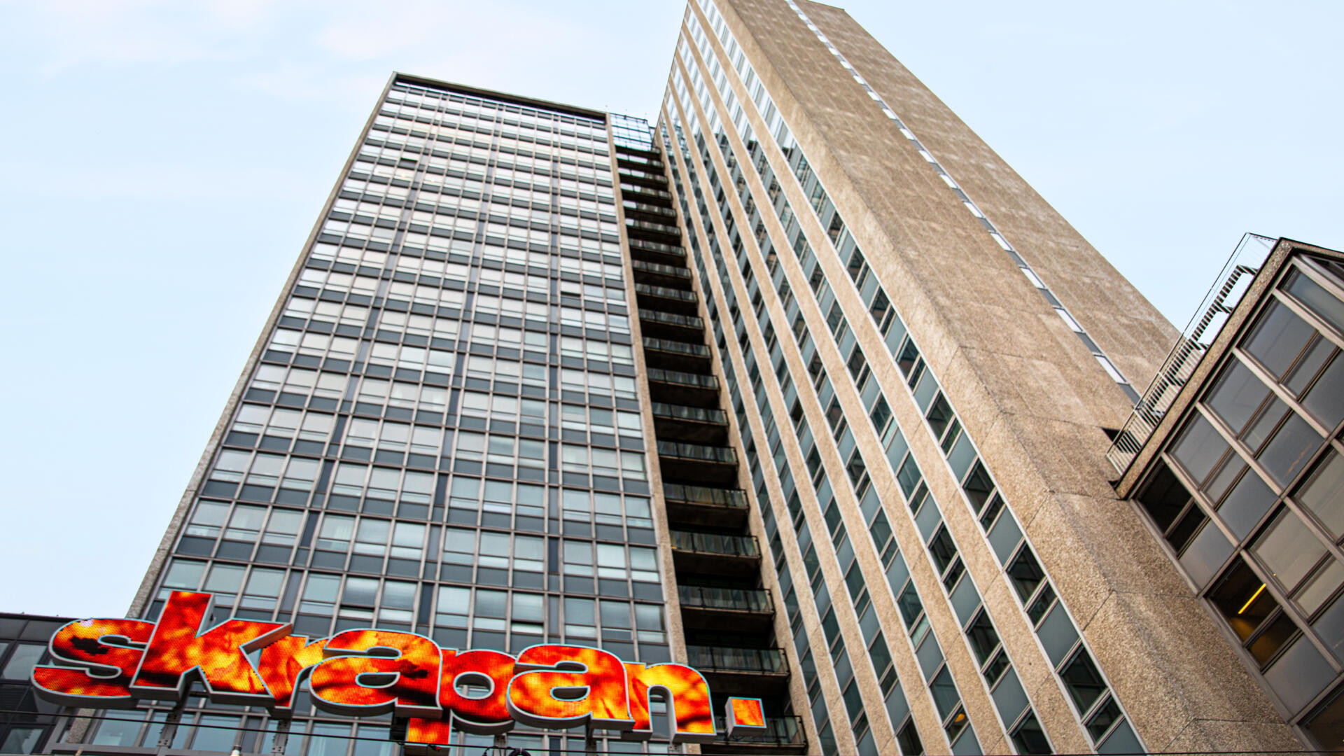 Den betong- och glasförsedda fasaden av gamla skatteskrapan sträcker sig upp mot en blå himmel. Längst ner syns den färgsprakande skylten som lyder "skrapan".